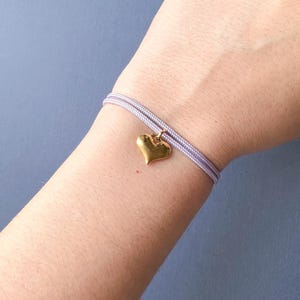 Può includere: Un braccialetto in cordoncino lavanda con un ciondolo a forma di cuore dorato.