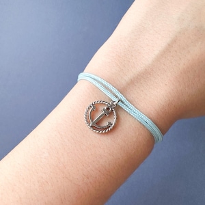 Puede incluir: Un brazalete de cordón azul claro con un dije de ancla de plata. El ancla está en un círculo con un diseño de cuerda.