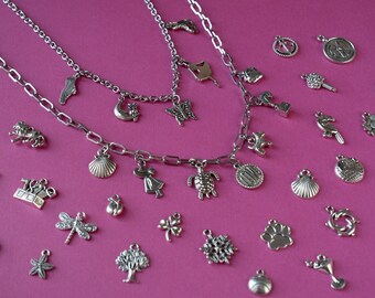 Può includere: Due catene d'argento con vari ciondoli, tra cui una conchiglia, una tartaruga, una libellula, un albero, un'impronta di zampa, un fiocco di neve, un trifoglio, un bicchiere da martini, un girasole, un uccello, una chiave, una corona, una luna, una farfalla, un gatto, una scarpa, una ragazza, una conchiglia, un'impronta di zampa, un'ancora e una bussola. I ciondoli sono su uno sfondo rosa.