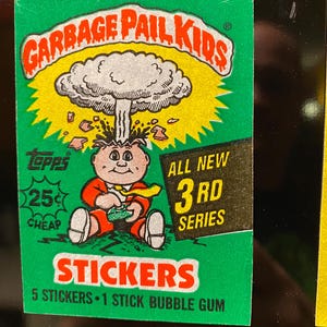Puede incluir: Una pegatina vintage de Garbage Pail Kids, con un personaje de dibujos animados con la cabeza explotando. La pegatina es verde con detalles en rojo y amarillo, y el texto "GARBAGE PAIL KIDS" se muestra de forma destacada. Dice "ALL NEW 3RD SERIES" y "STICKERS".