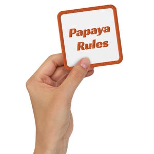 Puede incluir: Una mano sosteniendo un parche cuadrado blanco con un borde naranja. El parche tiene el texto "Papaya Rules" en bordado naranja.