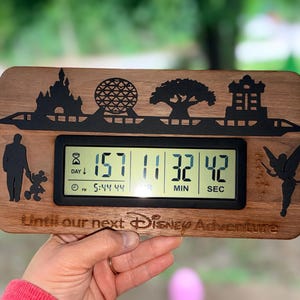 May include: A rectangular wooden countdown clock featuring a black silhouette of a monorail, castle, geosphere, tree, and tower. The digital display shows 157 days, 11 hours, 32 minutes, and 42 seconds. The text reads, Until our next Disney Adventure.
