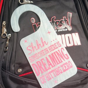 May include: A light blue door hanger with the text "Shhh... SOMEONE IN HERE IS DREAMING OF HITTING ZERO" in pink. The hanger is resting on a black backpack with red trim and the logo "iDance Fest!"