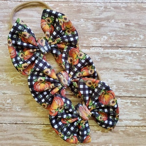 May include: Three black and white gingham hair bows with a pumpkin and sunflower print. The bows are tied together with a brown elastic headband.