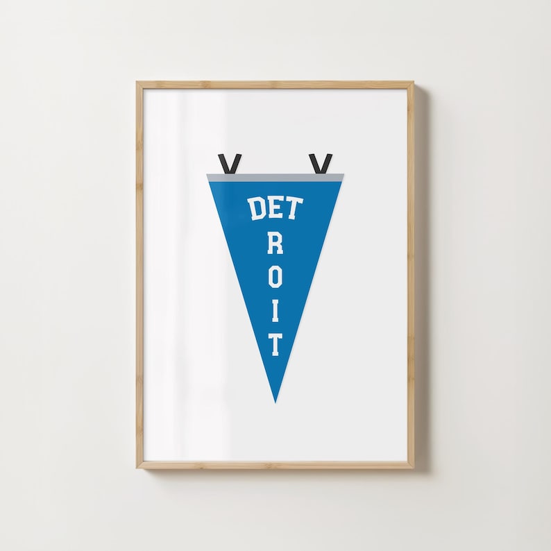 May include: A framed print featuring a blue pennant with the word "DETROIT" in white block letters. The pennant has a gray top and black accents. The frame is a light wood color.