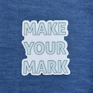 May include: A white sticker with the text "MAKE YOUR MARK" in light blue letters with a black outline. The sticker is on a blue fabric background.