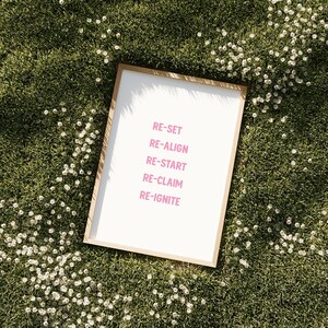 May include: A framed print with the words "RE-SET, RE-ALIGN, RE-START, RE-CLAIM, RE-IGNITE" in pink lettering. The frame is light brown. The print is on a white background and is set on a bed of green grass with small white flowers.