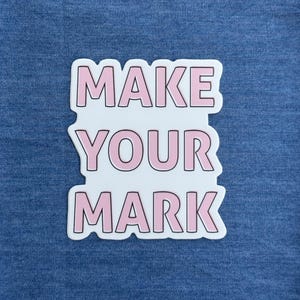 May include: A white sticker with the words "MAKE YOUR MARK" in pink block letters. The sticker has a white border and is set against a blue denim background. The text is stacked vertically.