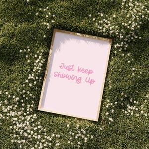 Könnte beinhalten: Ein gerahmter Druck mit dem Text "Just Keep Showing Up" in rosa Schrift. Der Druck hat einen hellrosa Hintergrund und einen goldfarbenen Rahmen. Der Druck liegt auf einem Bett aus grünem Gras mit kleinen weißen Blumen.