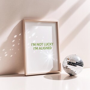 Puede incluir: Un cuadro enmarcado con el texto "I'M NOT LUCKY I'M ALIGNED" en letras verdes. Una bola de discoteca plateada se encuentra a la derecha del marco. El marco es de madera clara.