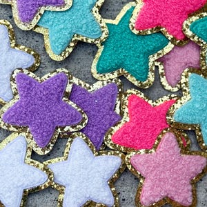 May include: A collection of colorful star-shaped embellishments. The stars are in shades of pink, purple, blue, and teal, with a textured surface and gold glitter trim. These decorative stars are ideal for crafts and DIY projects.