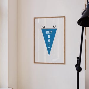 May include: A framed print featuring a blue pennant with the word "DETROIT" in white capital letters. The pennant is framed in a light brown wooden frame and hangs on a white wall. Two black V shapes are at the top of the pennant.