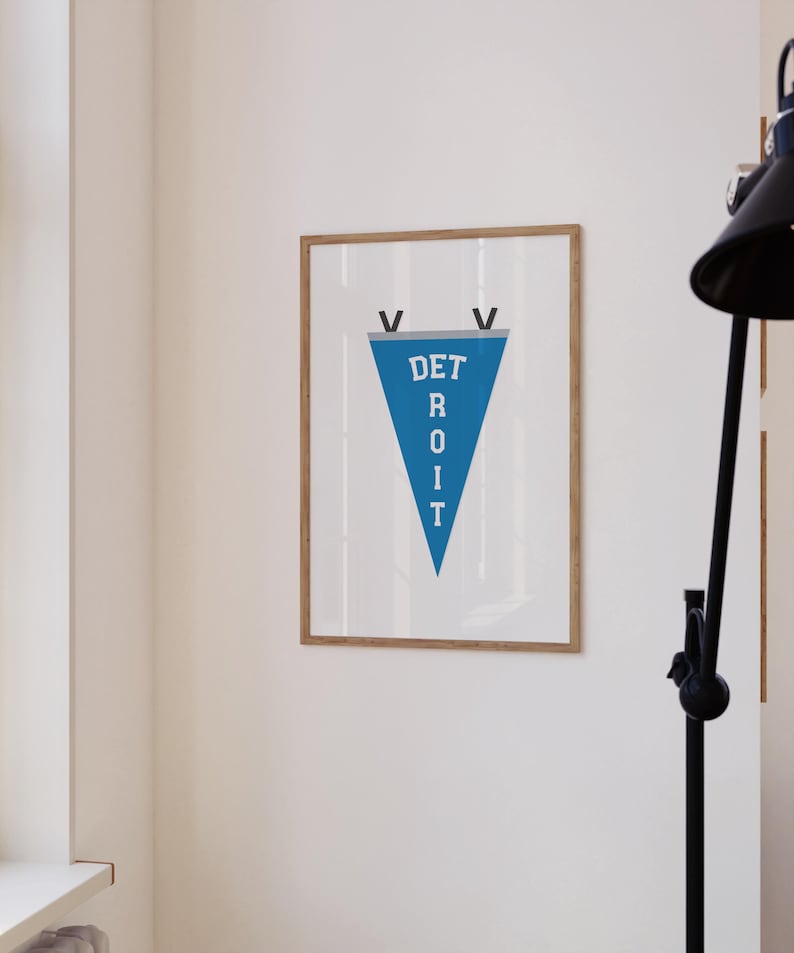 May include: A framed print featuring a blue pennant with the word "DETROIT" in white capital letters. The pennant is framed in a light brown wooden frame and hangs on a white wall. Two black V shapes are at the top of the pennant.