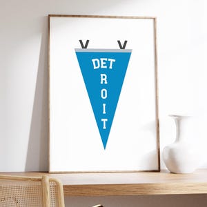 May include: A framed print featuring a blue pennant with the word "DETROIT" in white capital letters. The pennant has a gray border and black accents. The print is displayed on a wooden shelf with a white vase and a chair.