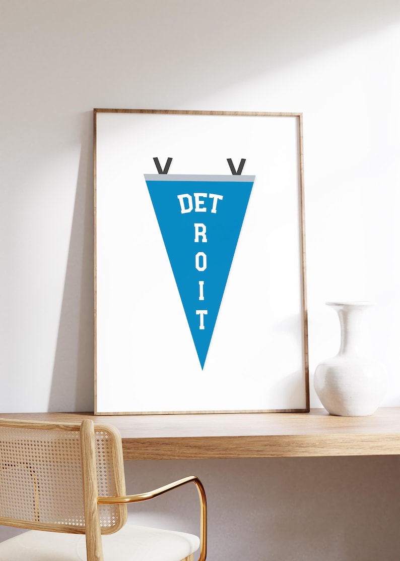 May include: A framed print featuring a blue pennant with the word "DETROIT" in white capital letters. The pennant has a gray border and black accents. The print is displayed on a wooden shelf with a white vase and a chair.