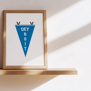 May include: A framed print featuring a blue pennant with the word "DETROIT" in white letters. The pennant is a triangle shape and is framed in a light brown wooden frame. The print is displayed on a wooden shelf against a white wall.