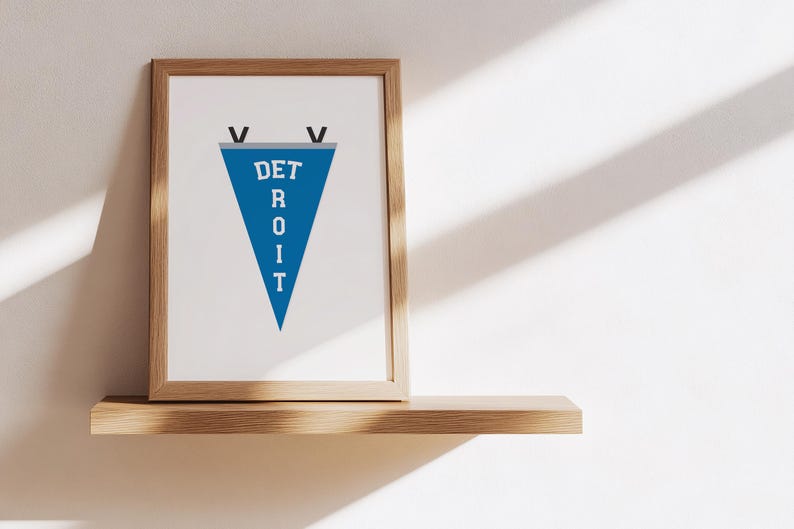 May include: A framed print featuring a blue pennant with the word "DETROIT" in white letters. The pennant is a triangle shape and is framed in a light brown wooden frame. The print is displayed on a wooden shelf against a white wall.