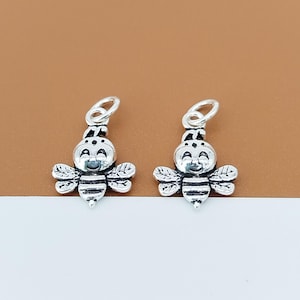 May include: Two silver-toned metal bee charms, each featuring a smiling face, striped body, and detailed wings. Each charm includes a small circular jump ring at the top for attachment, showcasing a polished finish with darkened recessed areas.