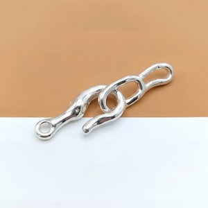 May include: A silver-colored metal clasp with a hook and eye design. The clasp has a textured, organic shape and is set against a white and tan background. The hook is curved, and the eye has a circular opening.