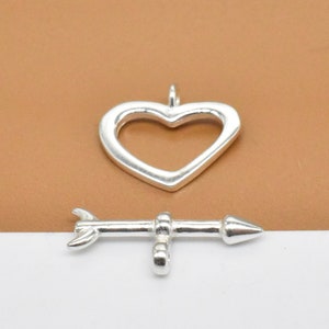 May include: Two silver charms, one shaped like a heart and the other like an arrow. The heart charm is open and the arrow charm has a pointed tip.