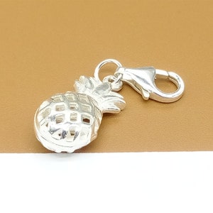 May include: Silver charm in the shape of a pineapple with a lobster clasp. The pineapple is detailed with a textured surface and a leafy top.