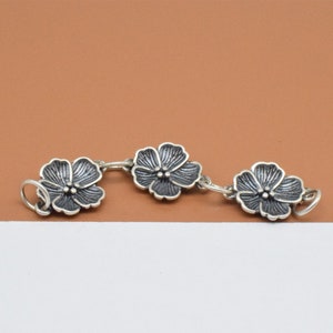 May include: A silver chain bracelet with three flower charms. The flowers are silver with a dark gray finish.