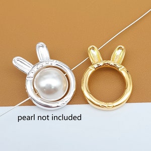 Op de afbeelding: Twee gouden ringen in de vorm van een konijn. Een ring heeft een ronde opening en de andere heeft een ronde opening met een witte parel in het midden. De tekst "parel niet inbegrepen" staat onder de ringen.