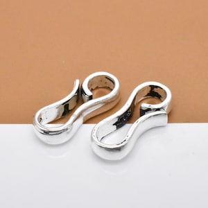 Könnte beinhalten: Zwei silberfarbene Metallverschlüsse, jeweils in Form eines liegenden 'S', werden vor einem braunen und weißen Hintergrund gezeigt.