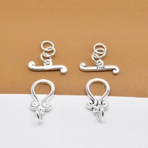 Könnte beinhalten: Zwei silberne Toggle-Verschlüsse mit einem dekorativen Design. Jeder Verschluss hat an einem Ende eine Schlaufe und am anderen Ende eine Stange. Die Stange hat an jeder Seite eine kleine Schlaufe. Die Verschlüsse sind mit "S925" auf der Stange gekennzeichnet.