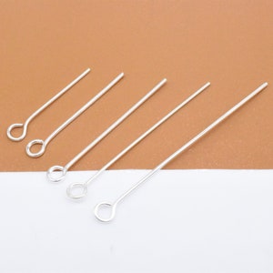 May include: Five silver head pins with a loop at one end. The pins are arranged in a row on a white surface.
