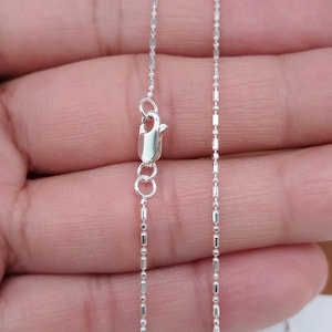 Puede incluir: Un delicado collar de plata con un pequeño cierre de mosquetón. La cadena está compuesta por pequeños eslabones rectangulares y ovalados alternos. El collar se muestra sobre un fondo neutro.