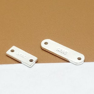 May include: Two silver-colored metal rectangular tags with rounded edges. The larger tag has the number "925" engraved on it. The smaller tag has the number "925" engraved on it.