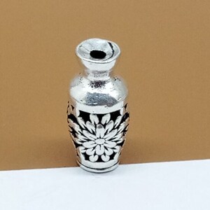 K&ouml;nnte beinhalten: Silberner Vasen-Anh&auml;nger mit floralem Design. Die Vase ist mit einem erhabenen Blumenmuster verziert und hat eine glatte, polierte Oberfl&auml;che.