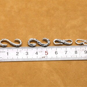 May include: Four silver-toned metal clasps with intricate designs, measured against a ruler marked in centimeters.