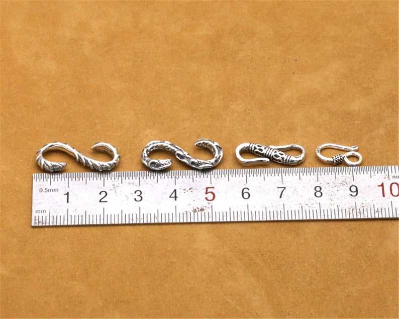 May include: Four silver-toned metal clasps with intricate designs, measured against a ruler marked in centimeters.