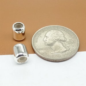 Op de afbeelding: Twee glanzende, zilverkleurige vatvormige kralen naast een Amerikaanse kwartdollar. De kralen hebben een cilindrische vorm met afgeronde uiteinden en een holle kern. De munt toont een profiel van George Washington.
