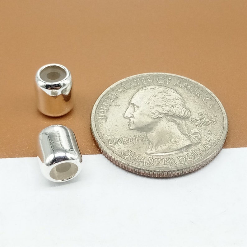 Op de afbeelding: Twee glanzende, zilverkleurige vatvormige kralen naast een Amerikaanse kwartdollar. De kralen hebben een cilindrische vorm met afgeronde uiteinden en een holle kern. De munt toont een profiel van George Washington.