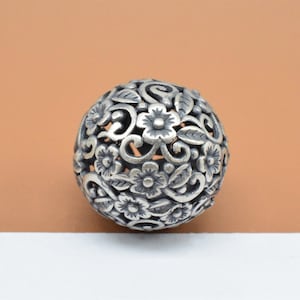 May include: A silver bead with a delicate floral design. The bead is round and has a hollow center, creating a delicate and intricate pattern.