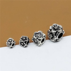 May include: Four silver charm beads with a floral design. The beads are different sizes and are arranged in a row from largest to smallest.