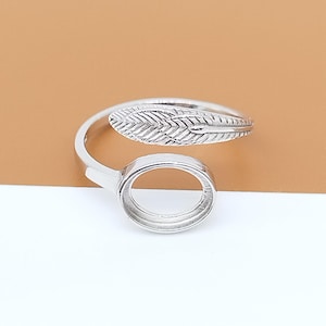 Puede incluir: Un anillo plateado con diseño de pluma y engaste circular. El detalle de la pluma es texturizado y el anillo es ajustable. El anillo se muestra sobre un fondo neutro.