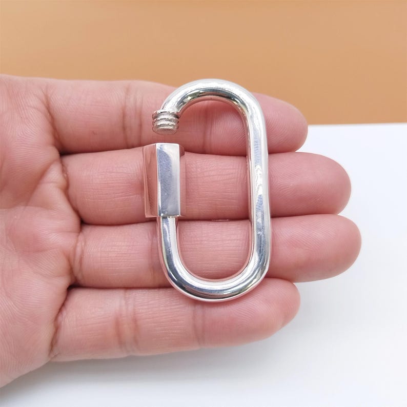 May include: A silver-colored, oblong carabiner with a screw-lock mechanism. The carabiner is open, revealing the threaded end of the locking mechanism. The background is a mix of white and tan.