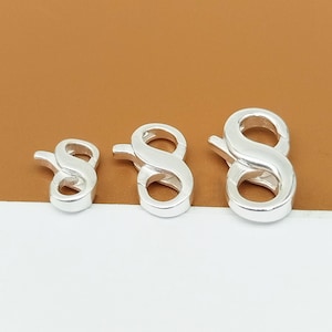 May include: Three silver-colored lobster clasps in varying sizes. Each clasp is shaped like the number eight, with a curved hook and a circular loop. The clasps are displayed against a brown and white background.