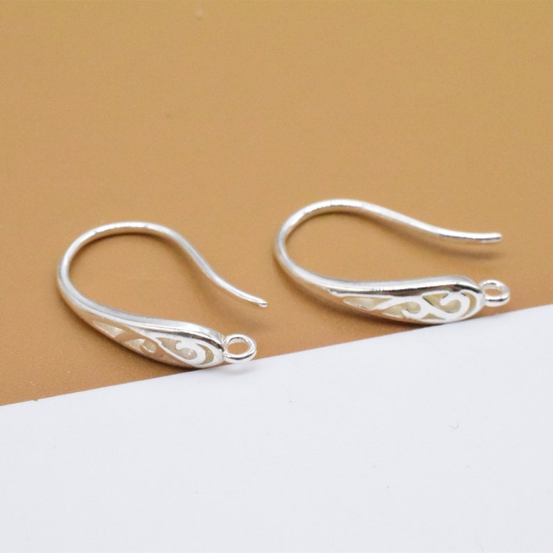 May include: Two silver earring hooks with a decorative, openwork design. The hooks are made of metal and have a smooth, polished finish.
