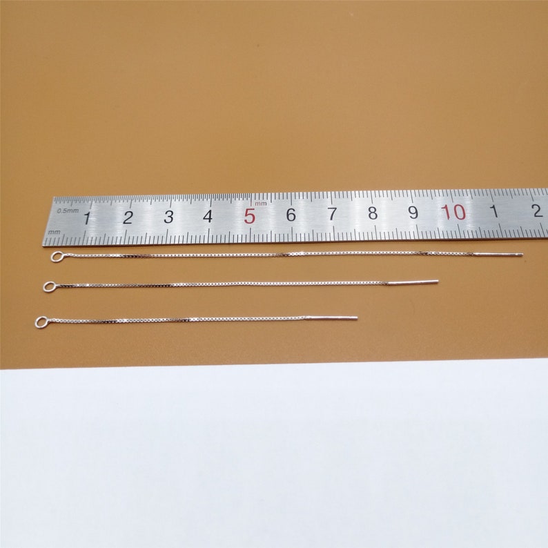 May include: Four silver chains of varying lengths, measured in centimeters, against a brown background. The chains are all the same width and have a delicate, box chain design.
