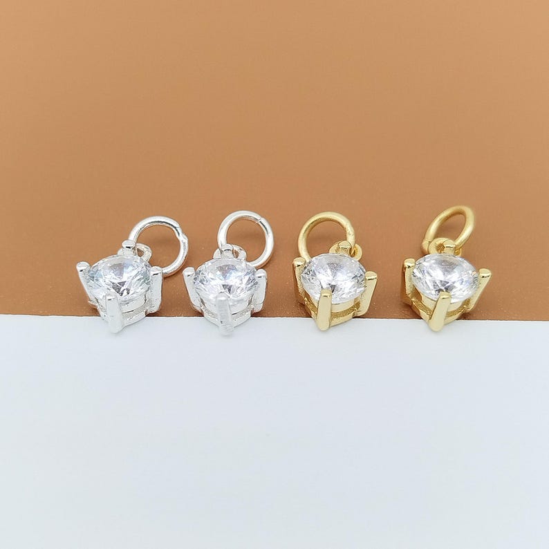 以下が含まれることがあります： 4つの小さなジュエリーチャーム。それぞれ4本爪の金属製バスケットにセットされた、透明なラウンドファセットカットストーンが特徴です。2つはシルバーカラー、2つはゴールドカラーで、上部には取り付け用の小さな丸いジャンプリングが付いています。
