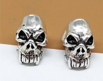 Puede incluir: Un par de pendientes de plata con forma de calavera. Las calaveras tienen detalles, incluyendo dientes y cuencas de los ojos.
