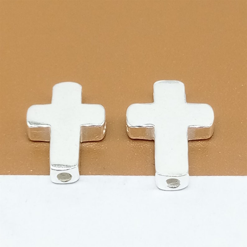 Op de afbeelding: Twee zilveren kruisbedels met een gat in het midden om aan een ketting of armband te rijgen.