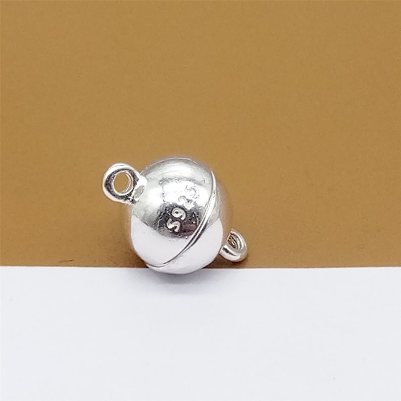May include: Silver magnetic clasp for jewelry making. The clasp is round and has a smooth, polished finish. The clasp is marked with "S925" indicating it is made of sterling silver.