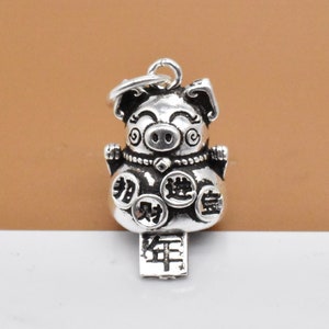 May include: Silver charm featuring a cartoon pig with coins and Chinese characters. The pig is wearing a necklace and has a happy expression. The charm is attached to a loop for hanging.