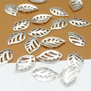 May include: A collection of silver-toned metal leaf charms with a cut-out design. The charms are scattered on a brown surface.